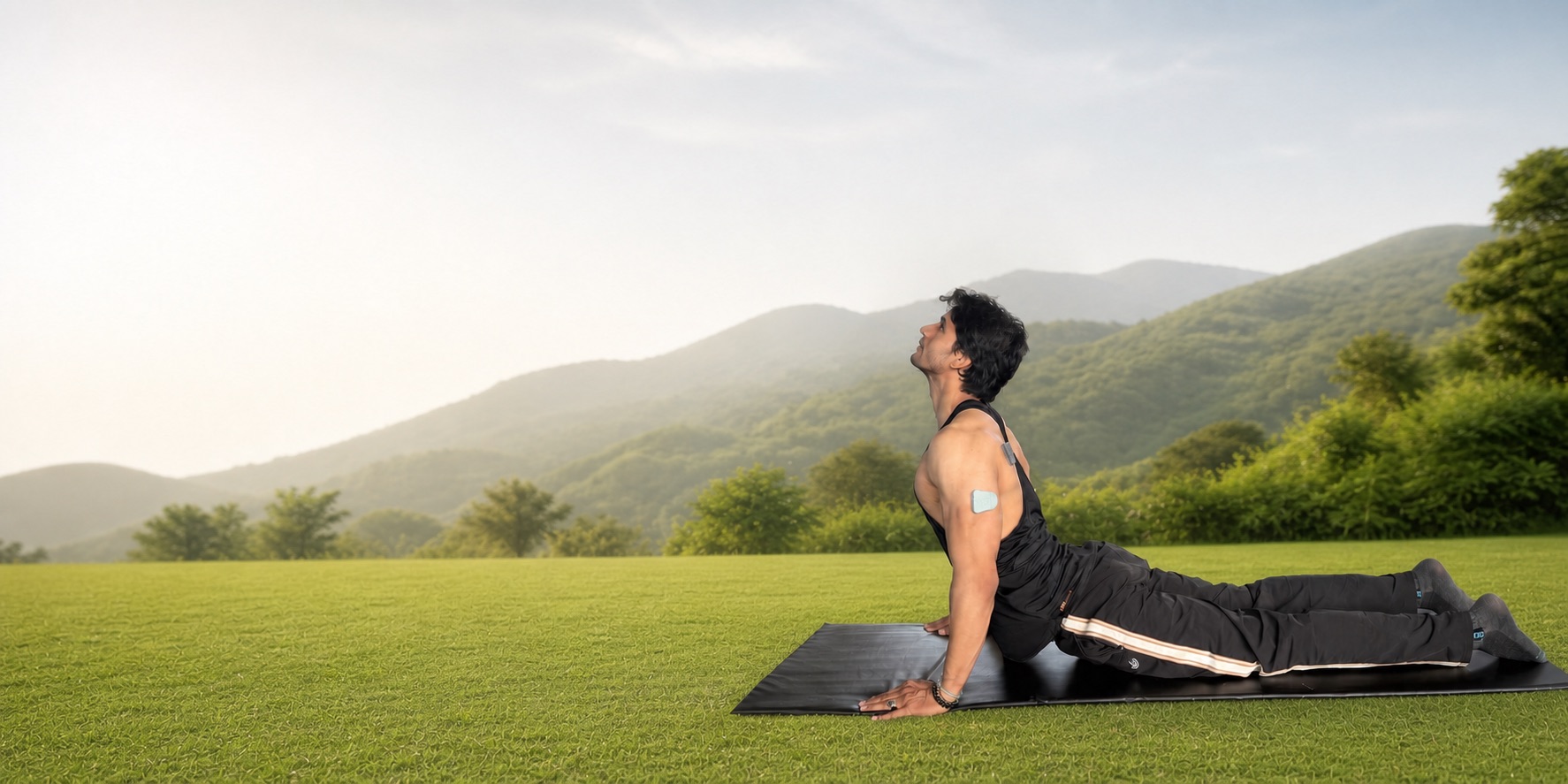 A man in a backbend yoga pose on a mat in a grassy field with mountains behind him, wearing a muscle sensor on his upper arm